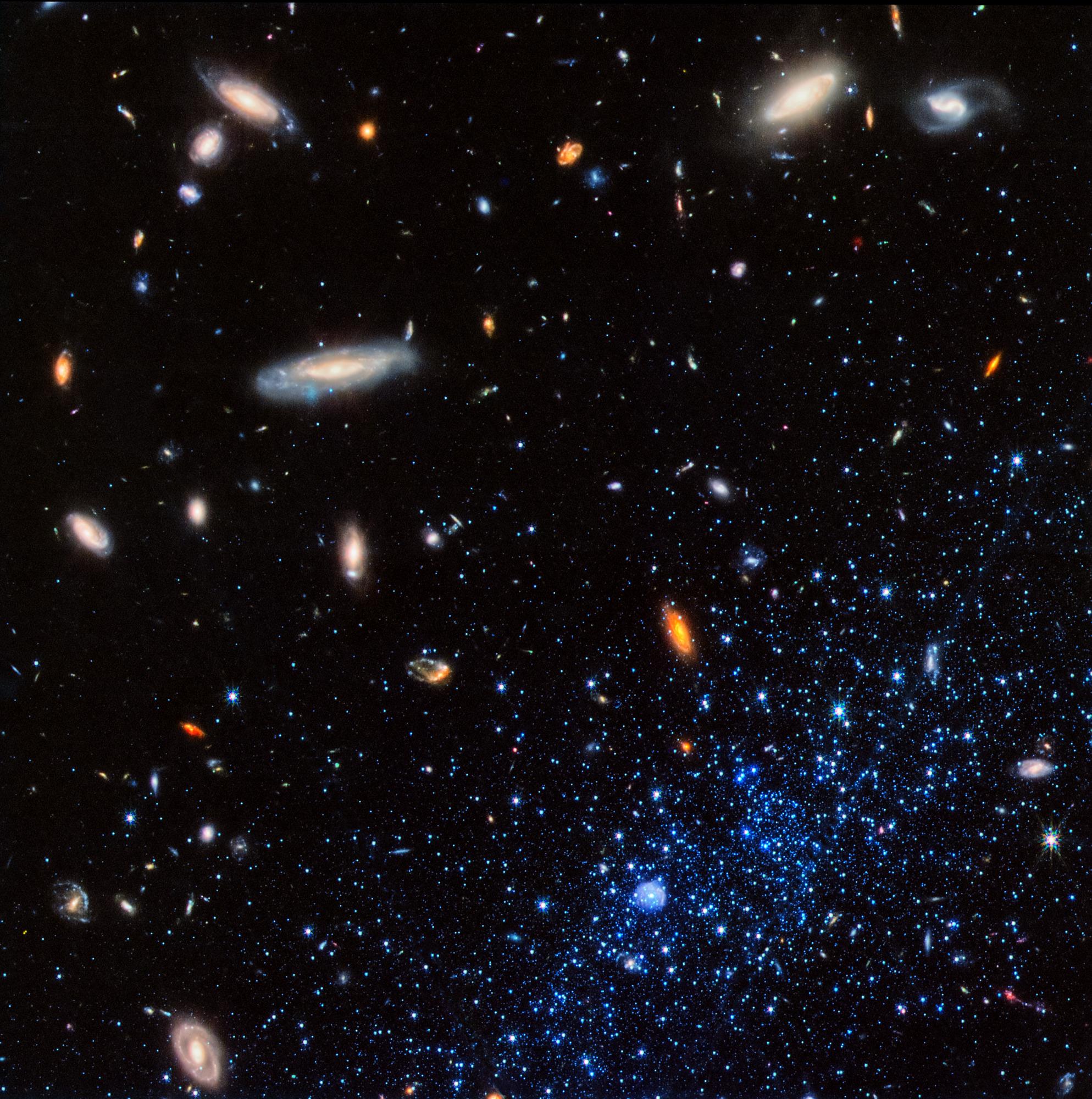 A concentration of bright blue stars occupies the bottom right corner of the image. At bottom center within them is a small blue bubble. The stars and bubble are part of a diffuse dwarf galaxy that extends beyond the image border. Background galaxies are <br />scattered across the image, with some particularly prominent spirals located at upper left and upper right. The background of space is black.